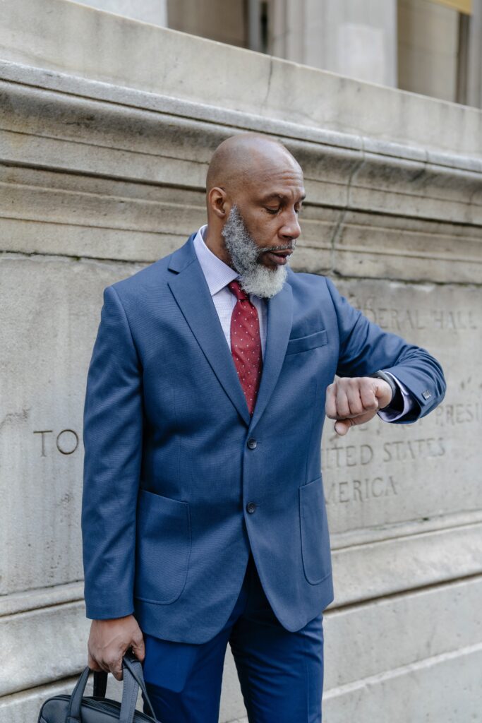 Image d'un homme noir au crâne rasé, en costume bleu et cravate rouge, regardant sa montre avec concentration, symbolisant la gestion du temps pour atteindre des objectifs.